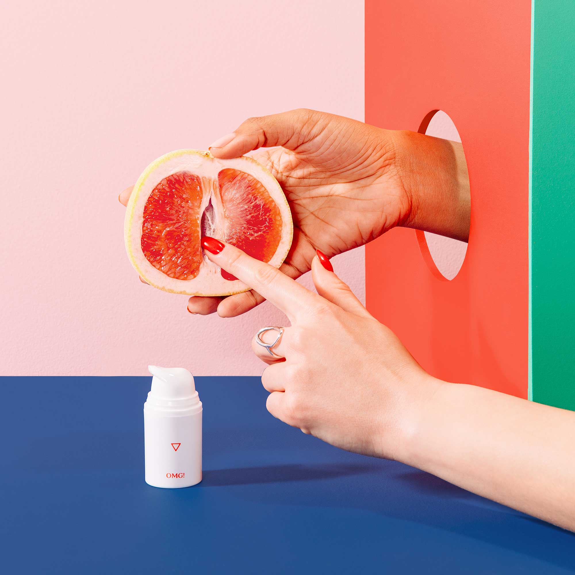 Hand holds a grapefruit while another hand touches it with one finger next to bottle of Wisp OMG! Cream on a blue surface, on a pink background