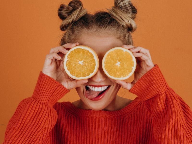 a woman in a red sweater sticking her tongue out and holding orange slices over her eyes, on an orange background