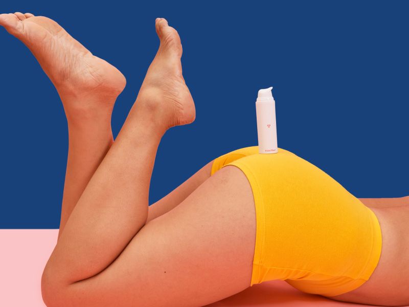 a bottle of Wisp's Even Out Hydroquinone Cream balancing on a woman's bottom in yellow shorts, on a blue and pink background