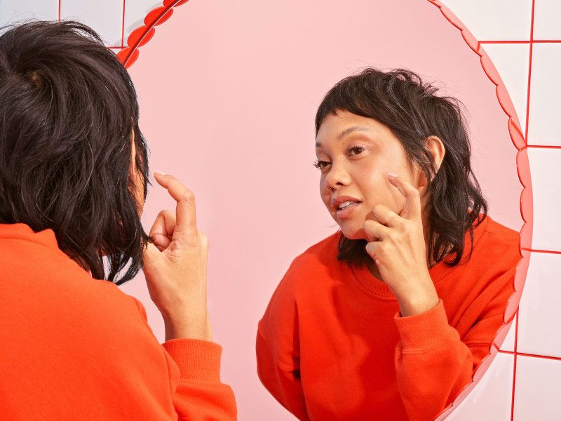 a woman in a red sweater inspecting her skin in a bathroom mirror