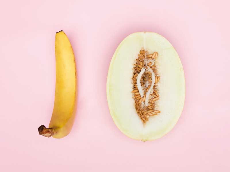A banana and a cut open vulva-shapes fruit on a pink surface