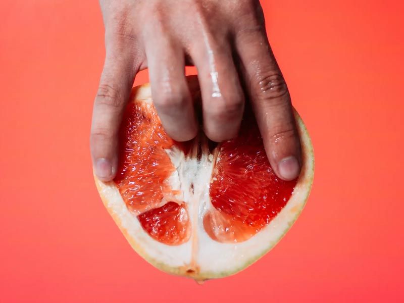 A hand suggestively holding a cut open grapefruit on a reddish background