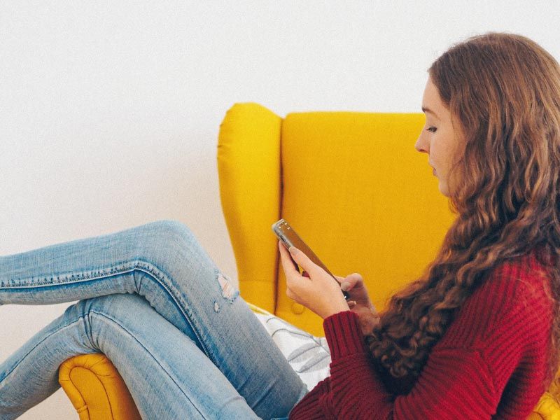 A female with brown hair wearing a red sweater, blue jeans and white shoes is sitting on a yellow chair looking up information on a mobile phone