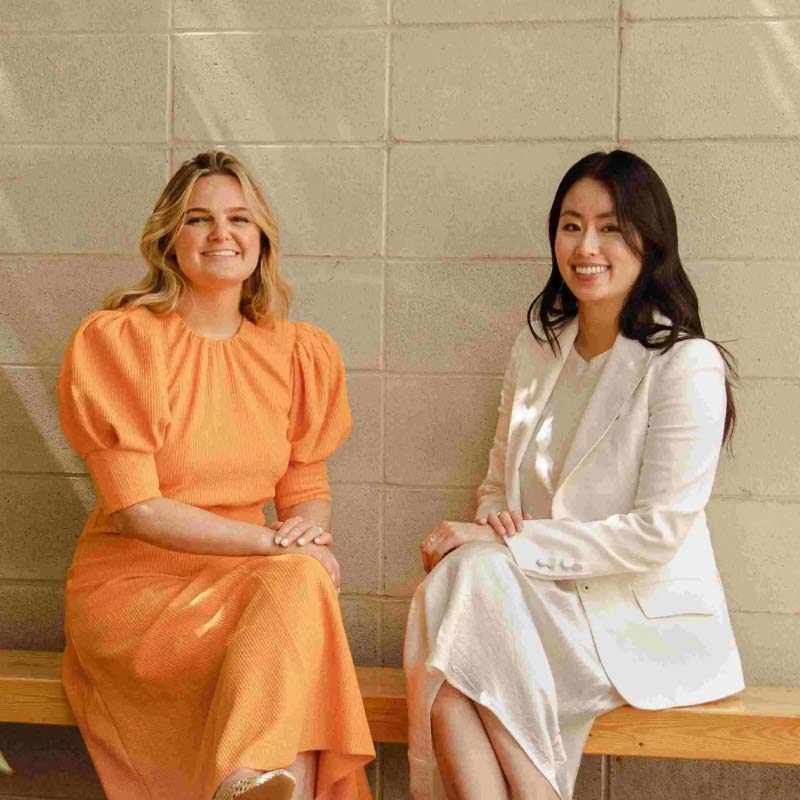 Stephanie Estey and Daphne Chen sitting on a wood bench with a neutral brick wall in the background