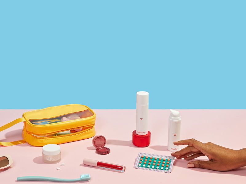 a woman's hand reaches for birth control and other Wisp medications next to a travel bag and toiletries on a pink and blue background