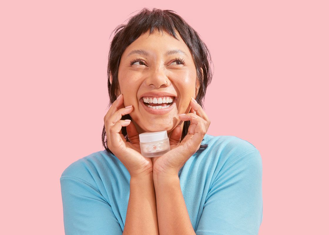 A female looking excited wearing a light blue shirt and holding a small glass Wisp pill jar in front of a pink background