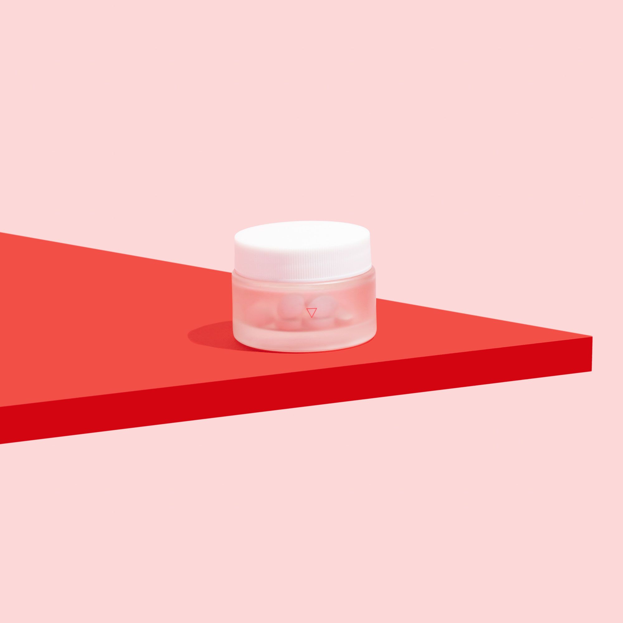 A small glass jar of antifungals to treat yeast infections on a pink background and red surface