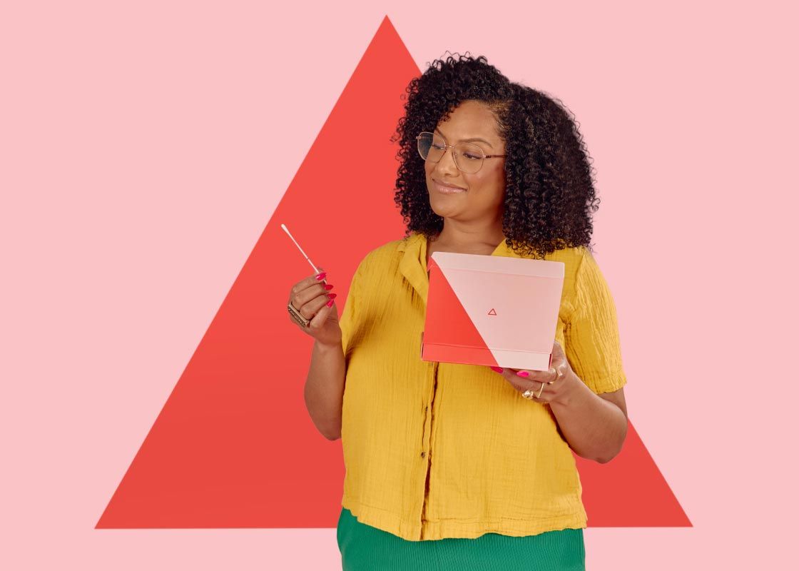 A smiling woman wearing a yellow shirt and green pants is holding a testing swab and Wisp diagnostics box in front of a pink and red geometric background
