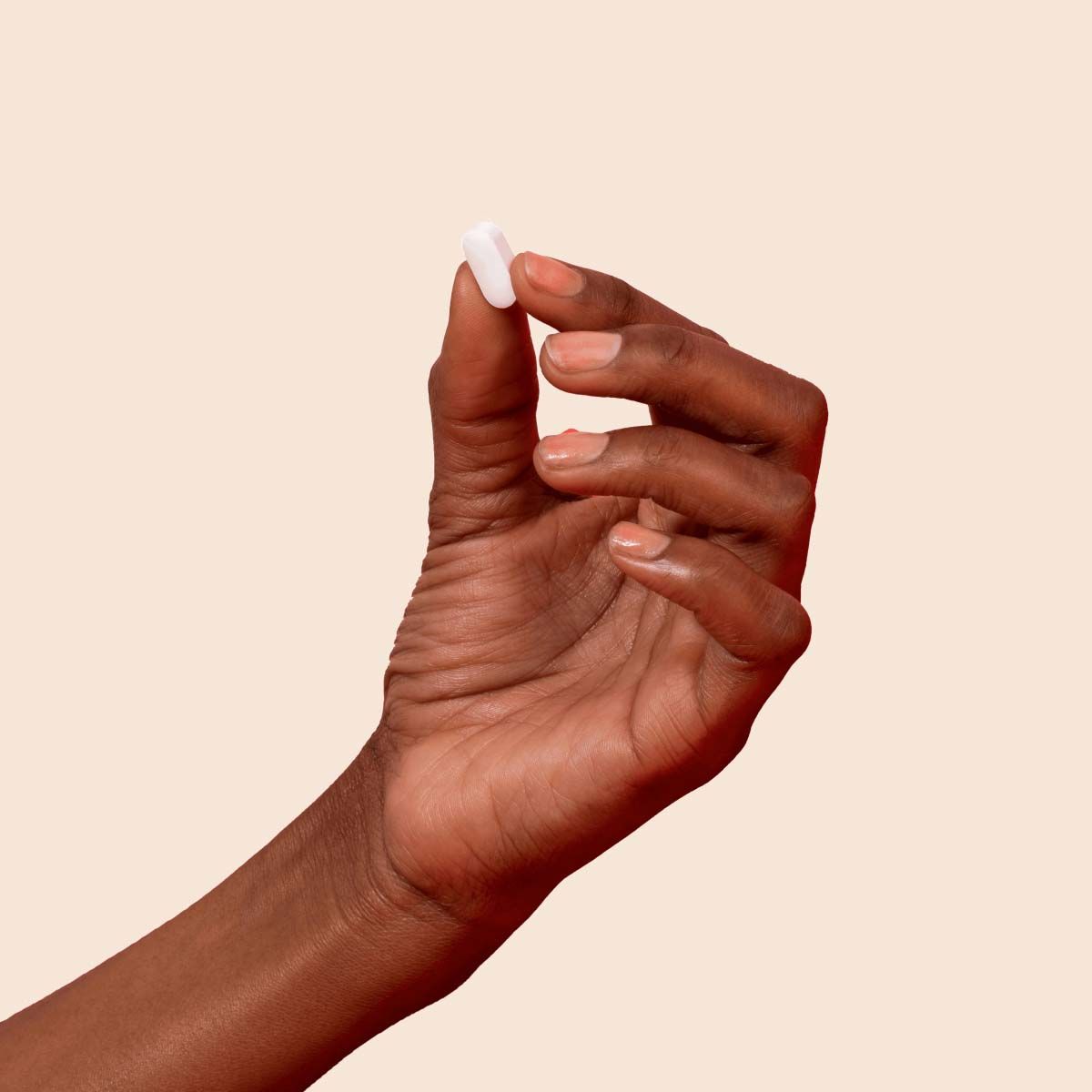 A close up of a woman's hand holding a white pill in front of a cream background
