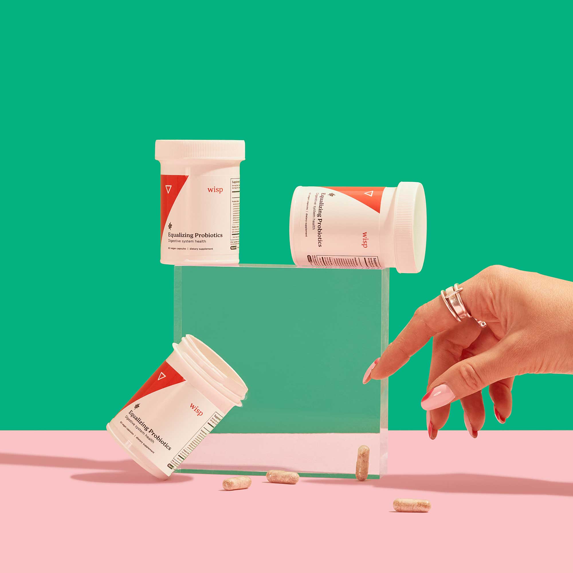 A woman's hand reaching for multiple bottles of Wisp Equalizing Probiotics with pills spilling out on a pink surface with a green background