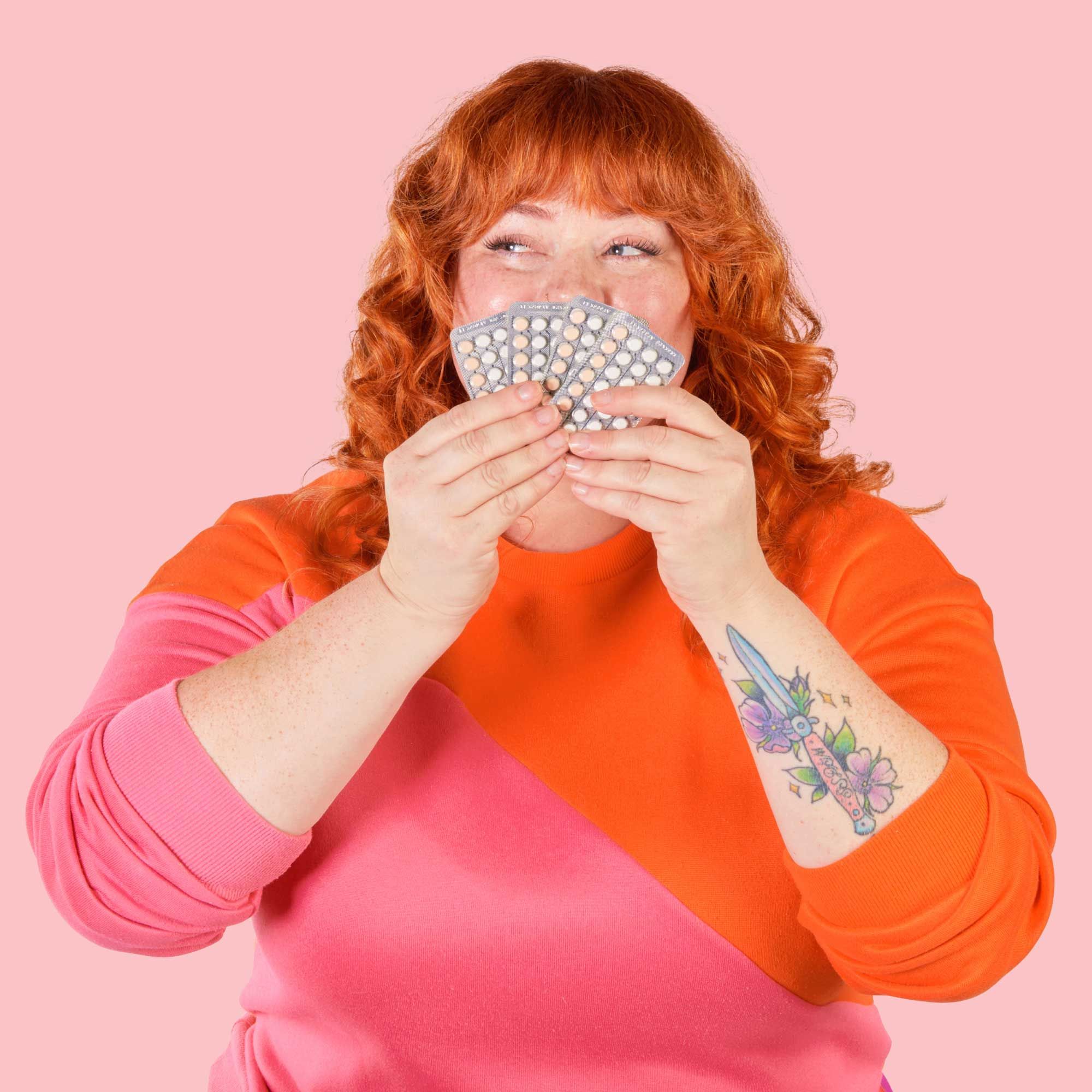 A woman with red curly hear wearing an orange and pink sweater is holding 4 birth control packets in front of a pink background