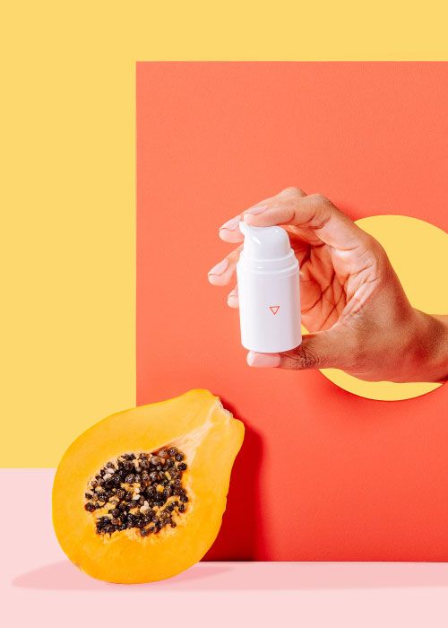 A woman's hand holding Wisp Calm Down through an orange cutout with a cut open papaya on a pink surface with a yellow background