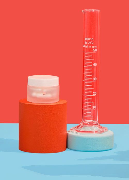 A small Wisp glass pill jar and a test tube balanced on colorful abstract shapes on a light blue surface with a red background