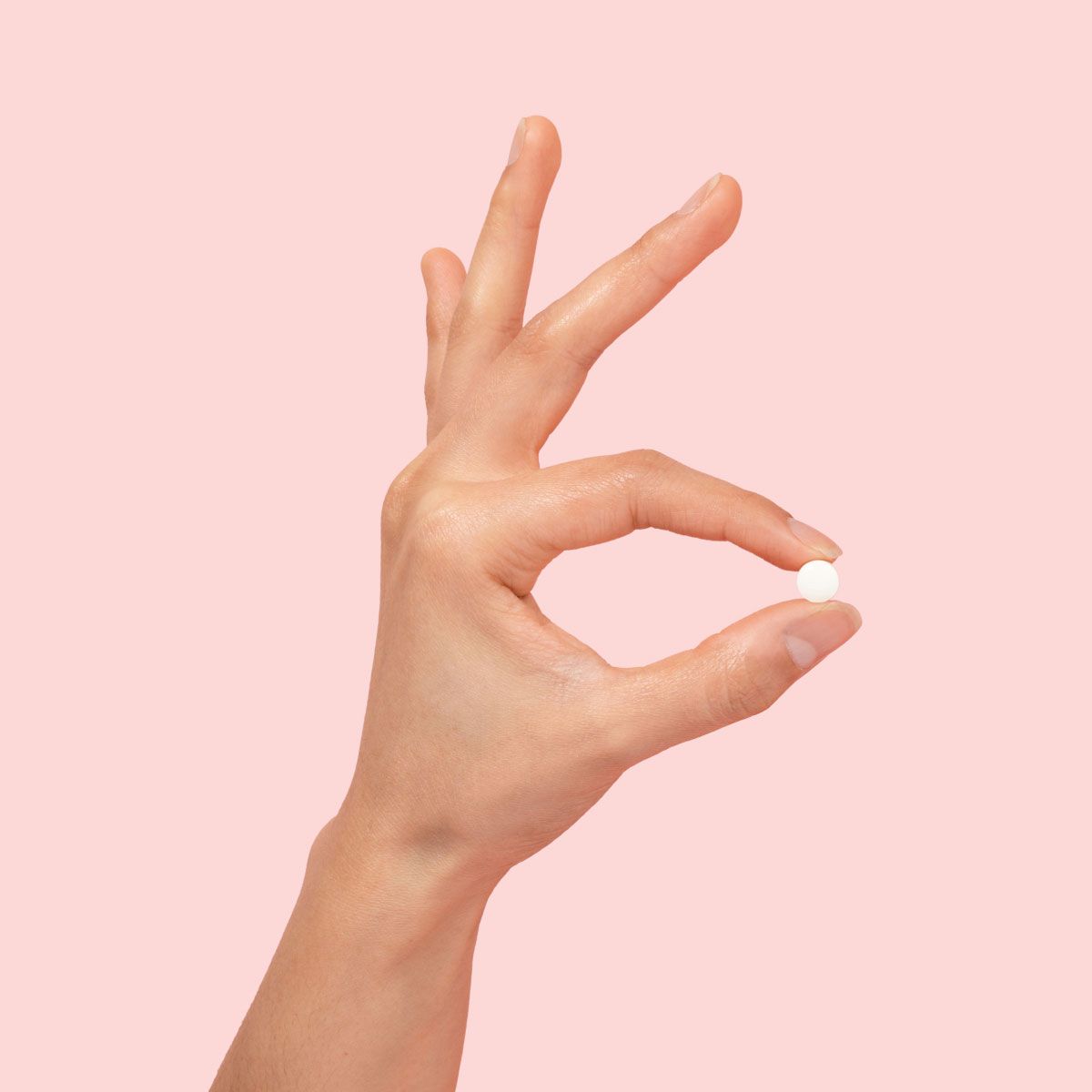 A person's hand holding a small round white pill between index finger and thumb in front of a pink background