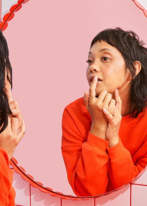 A woman wearing a red sweater is looking in the mirror while she applies a hydrocolloid patch to a cold sore on her lip