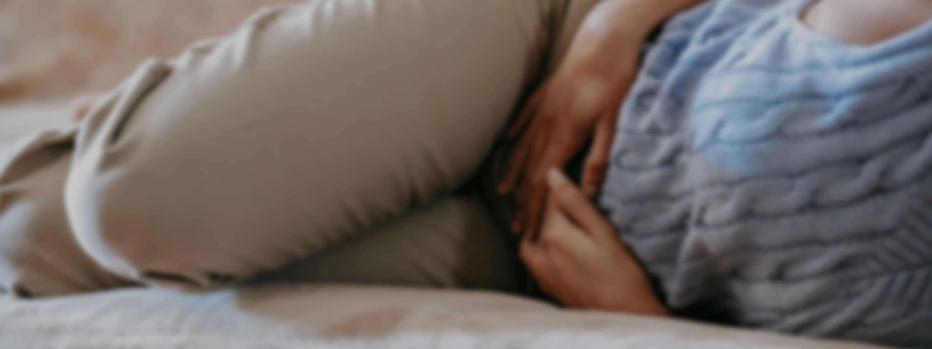 A woman wearing a blue sweater and tan pants is laying on a sofa with her hands on her abdomen