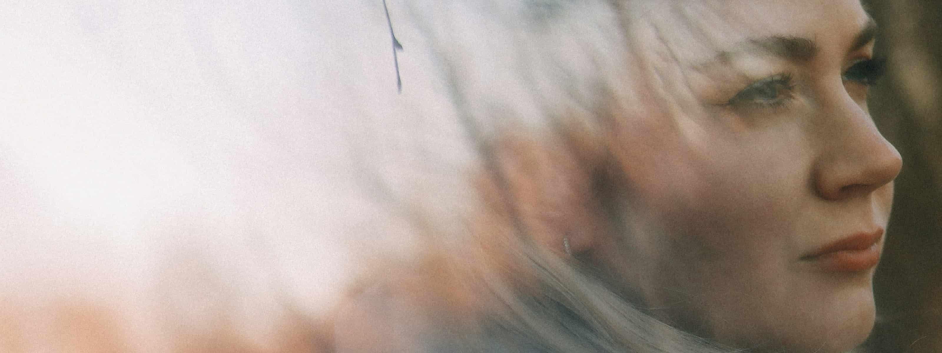 A woman gazing thoughtfully into the distance, with a soft double-exposure effect blending her face with a blurred natural background.