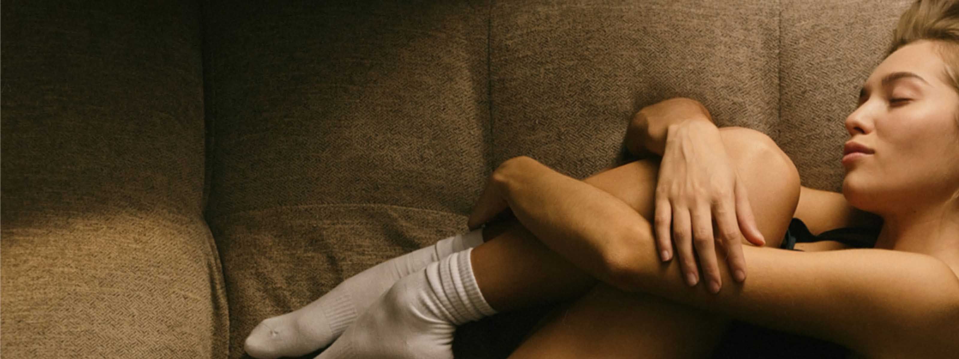 A person curled up on a couch, resting with eyes closed and arms wrapped around their legs.