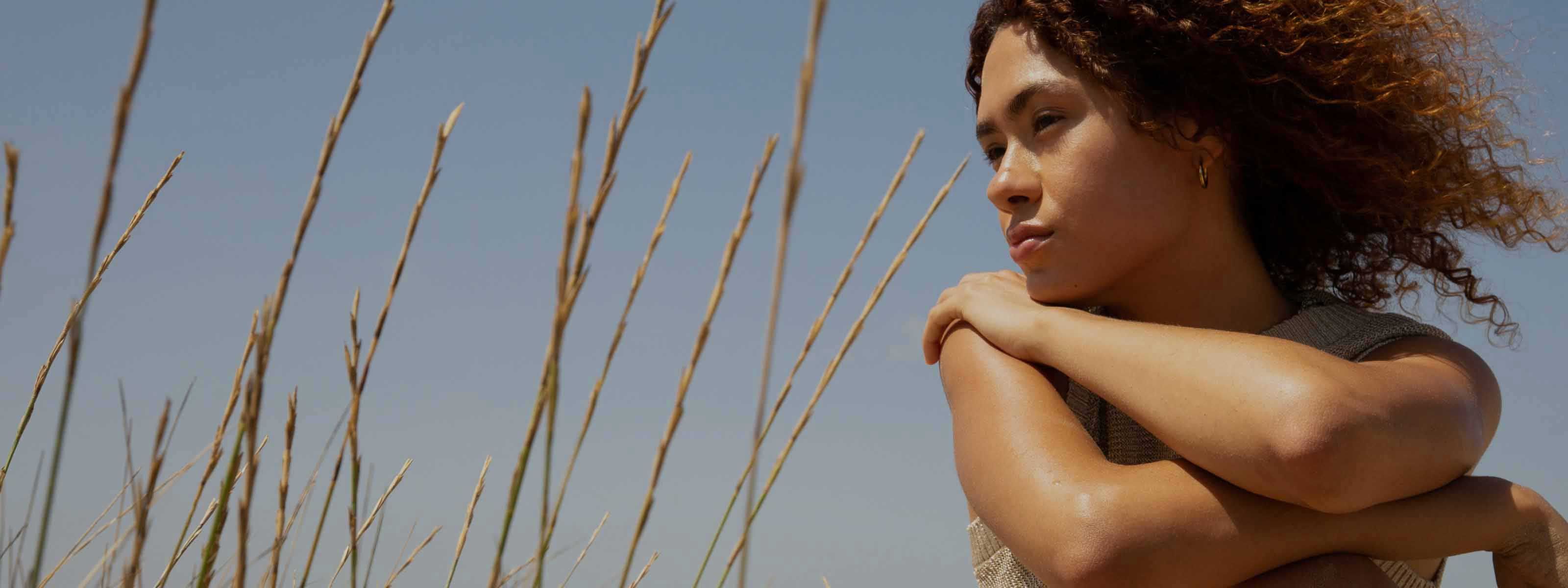 Woman with curly hair looking into the distance outdoors, with tall grass and a blue sky behind her.
