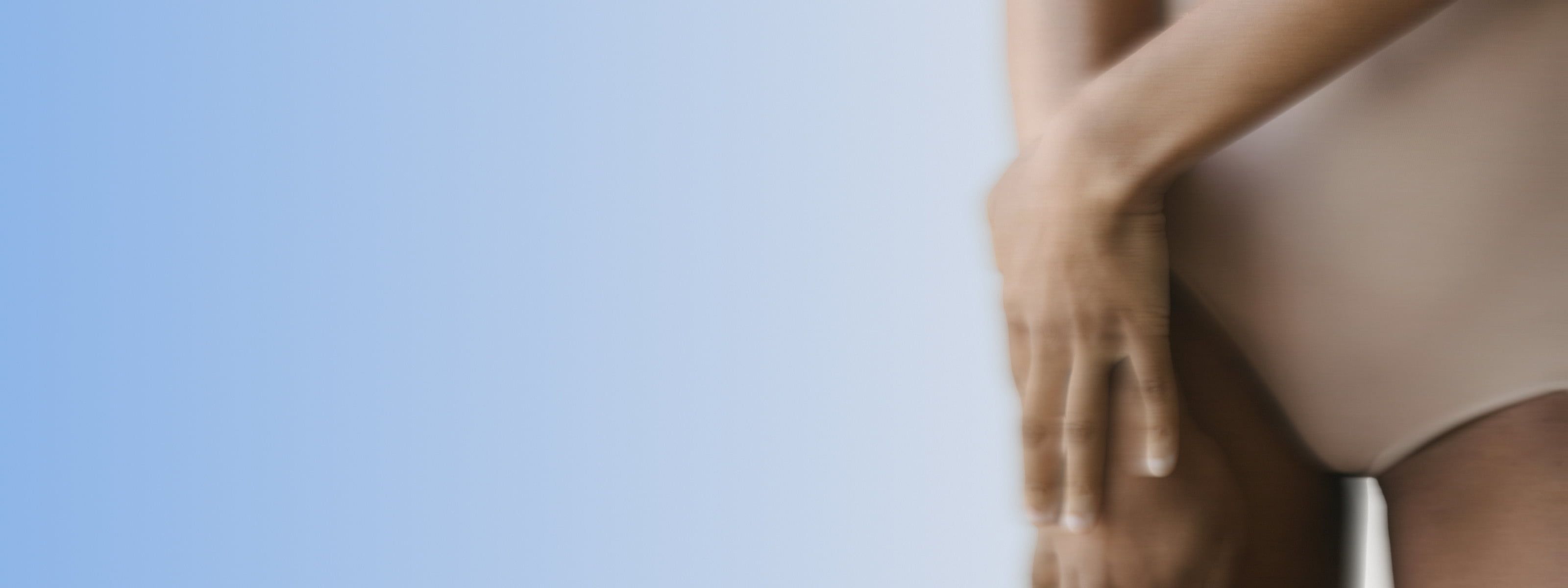 A person in nude-toned underwear with hands clasped over their lower abdomen, against a soft blue background.