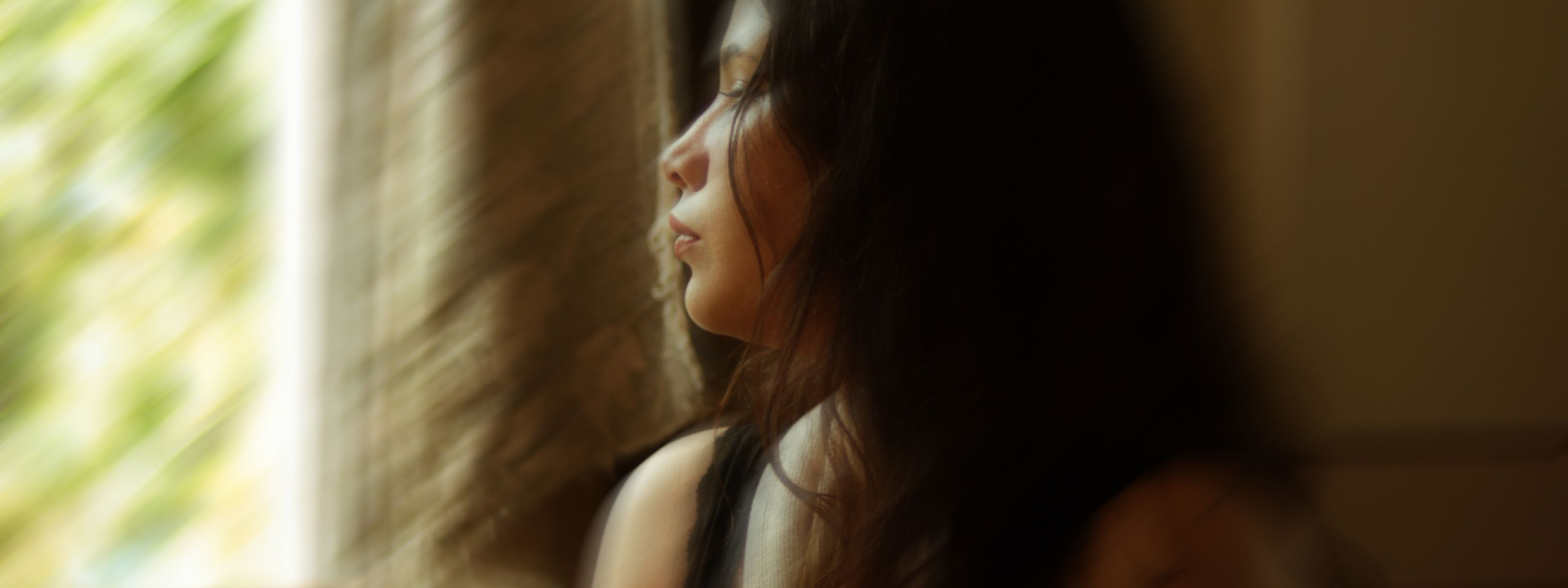 A woman with long dark hair in profile, gazing thoughtfully out a sunlit window with woven blinds, warm light illuminating her face against a softly blurred green outdoor backdrop.