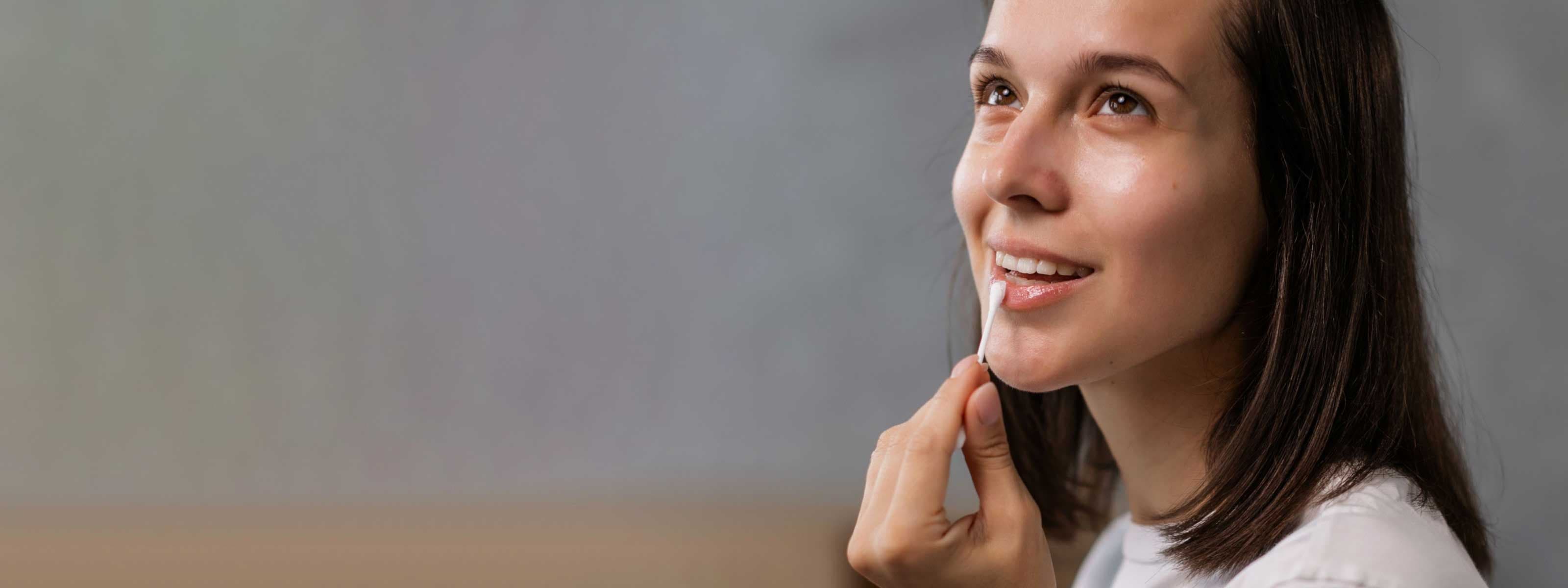 A smiling woman wearing a white shirt is applying a cream to her lip with a q tip