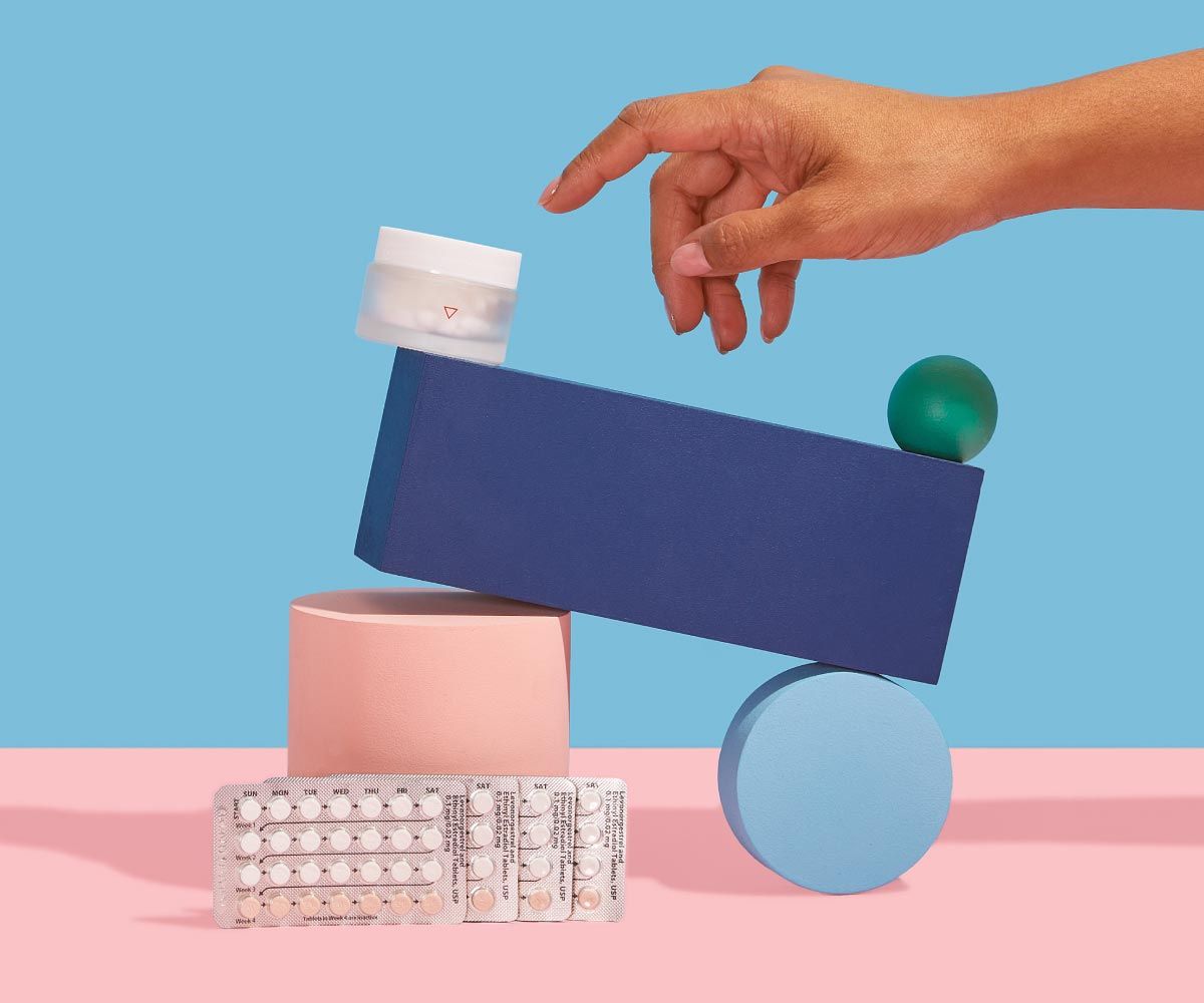 A Wisp glass pill jar and 4 birth control packets are balanced on colorful abstract shapes on a pink surface with a blue background