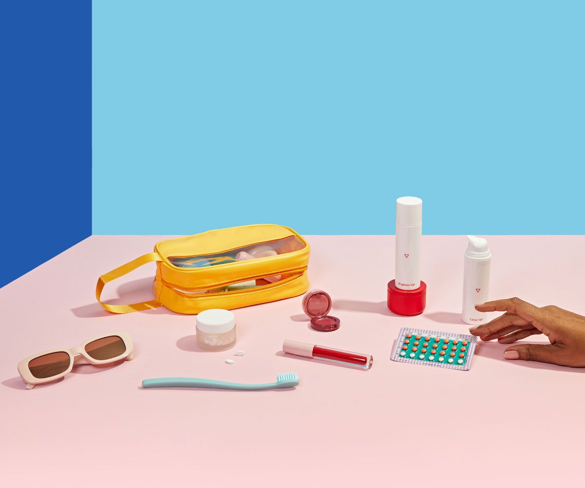 Various Wisp products, a yellow travel bag, sunglasses, a toothbrush, lip gloss and a birth control packet are laid out on a pink surface with a blue background