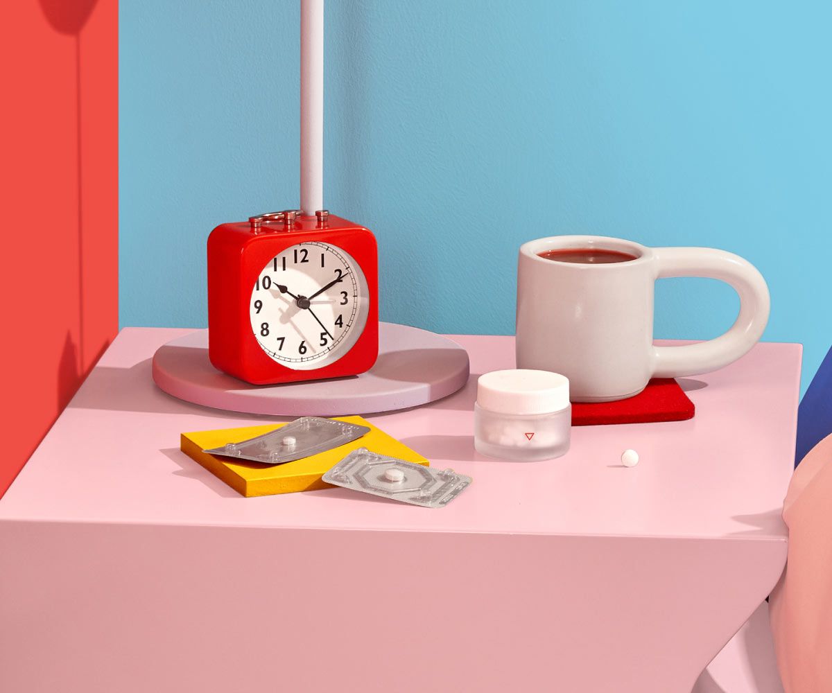 A Wisp glass pill jar and 2 emergency contraception packets are on a pink nightstand with a red alarm clock and white mug next to a bed with colorful bedding