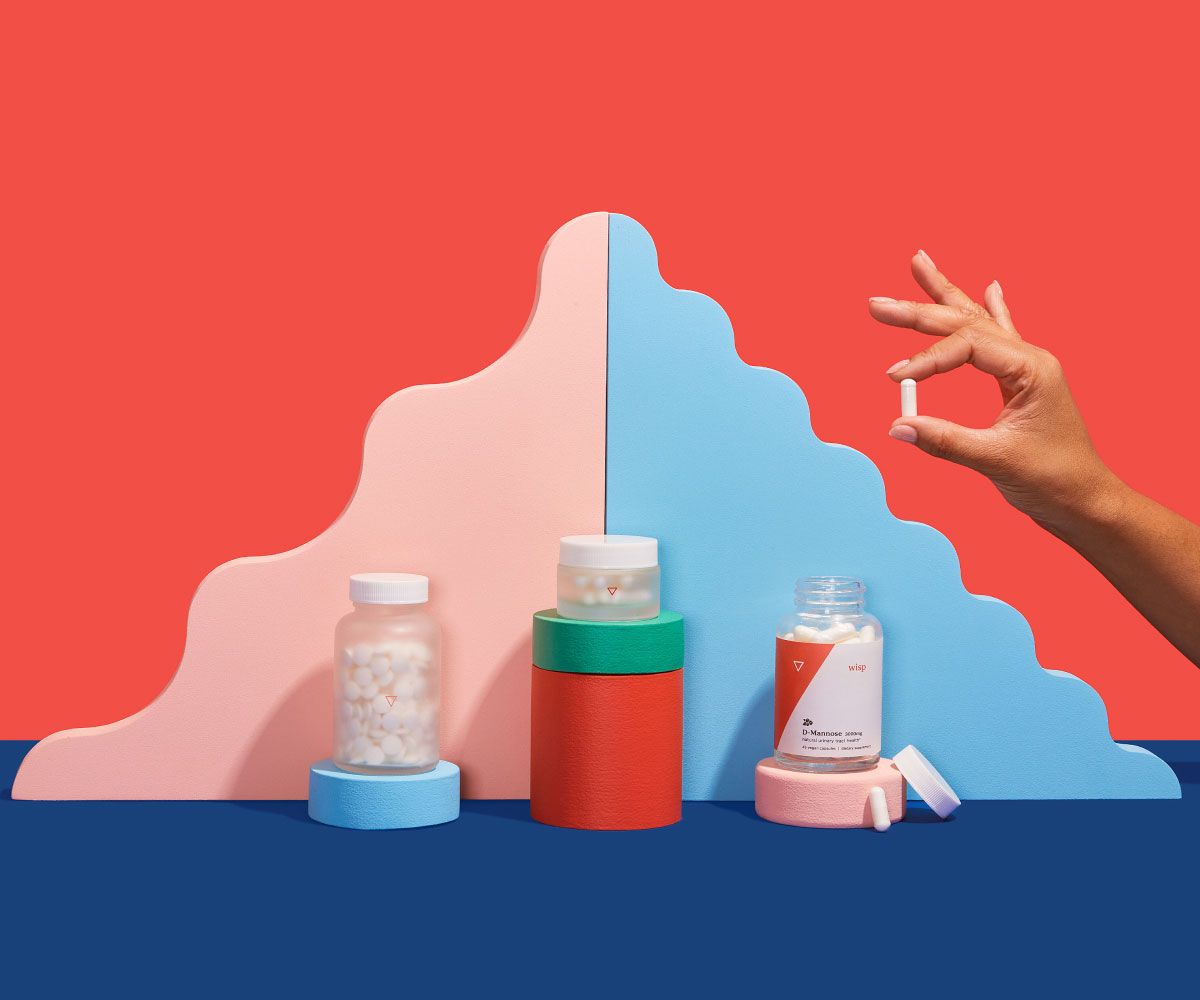 A person's hand is holding a pill next to various Wisp products balanced on colorful abstract shapes on a blue surface with a red background