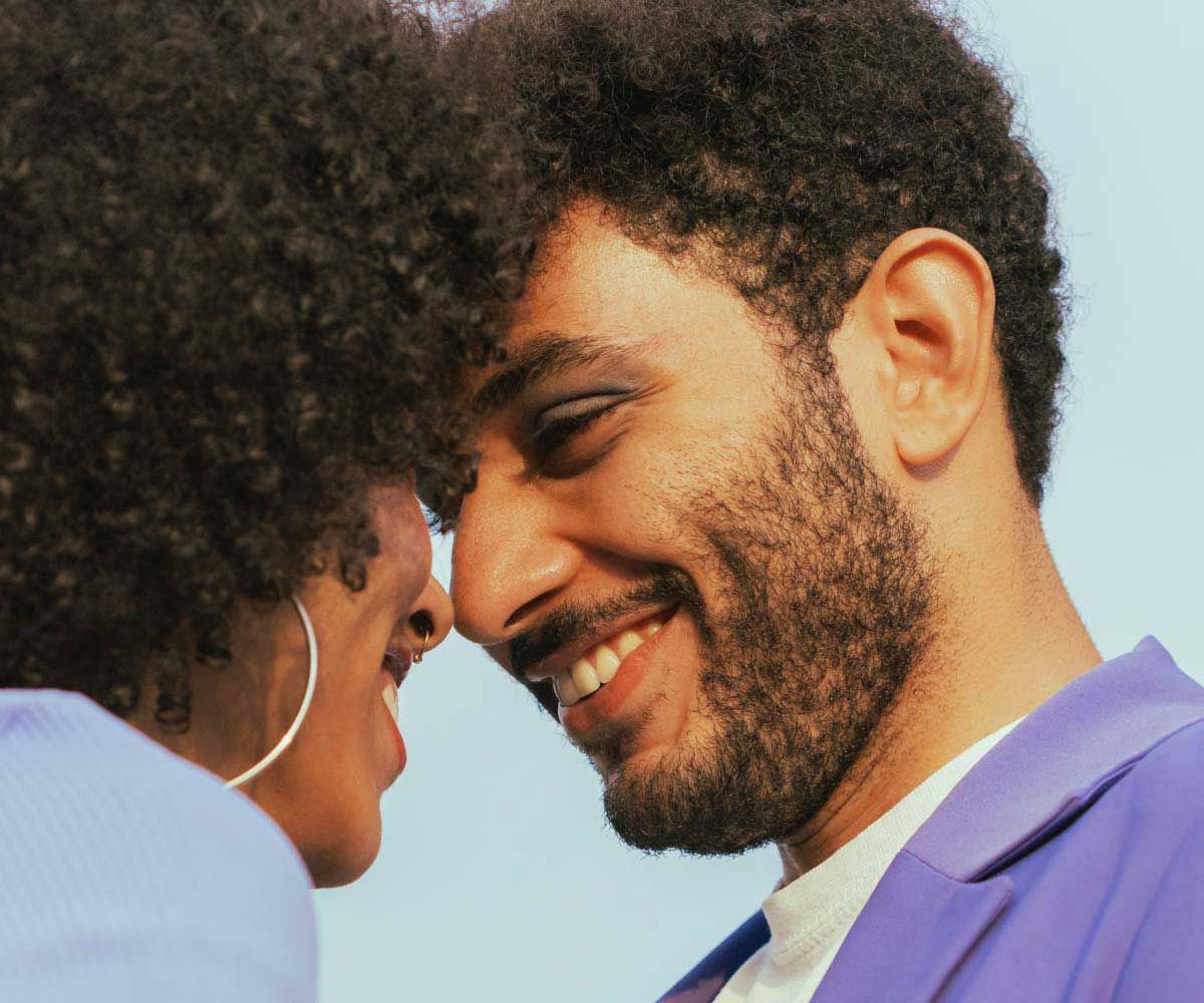 A woman and man with black curly hair and colorful clothing are looking into each other's eyes while smiling