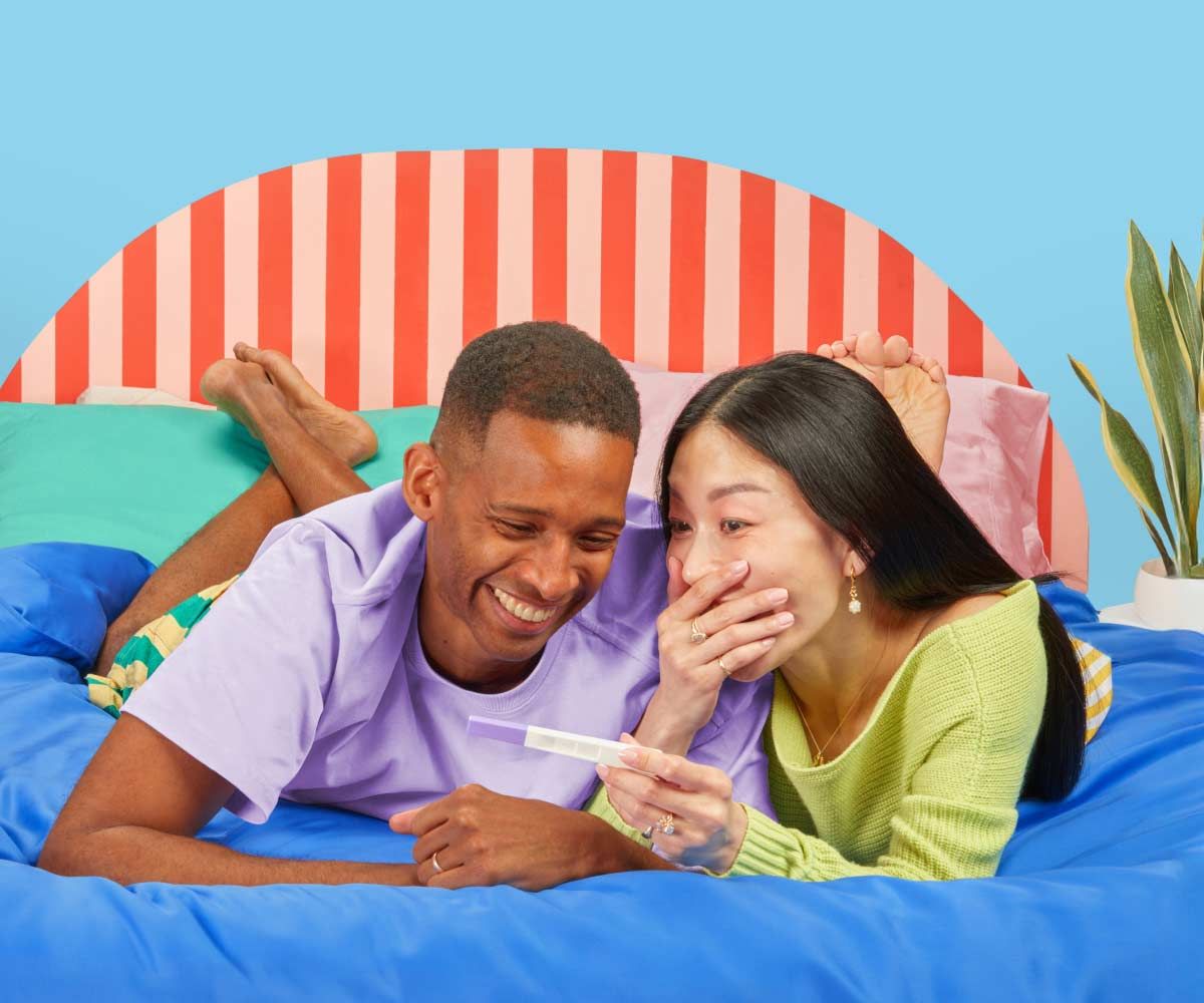 A man and woman wearing bright colored clothes are laying on a bed and smiling at each other while holding a pregnancy test