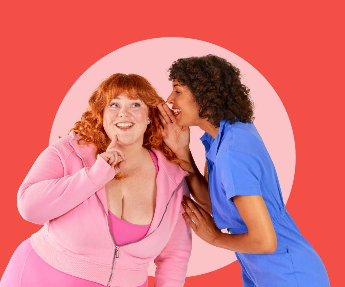 A woman wearing blue is whispering into another woman's ear who is smiling and wearing pink clothing