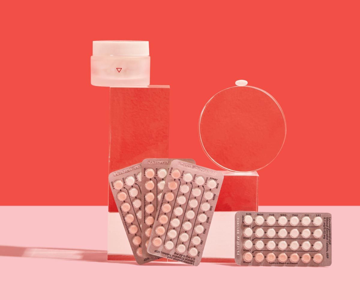 4 birth control packets and a small glass pill jar are balanced on clear abstract shapes on a pink surface with a red background