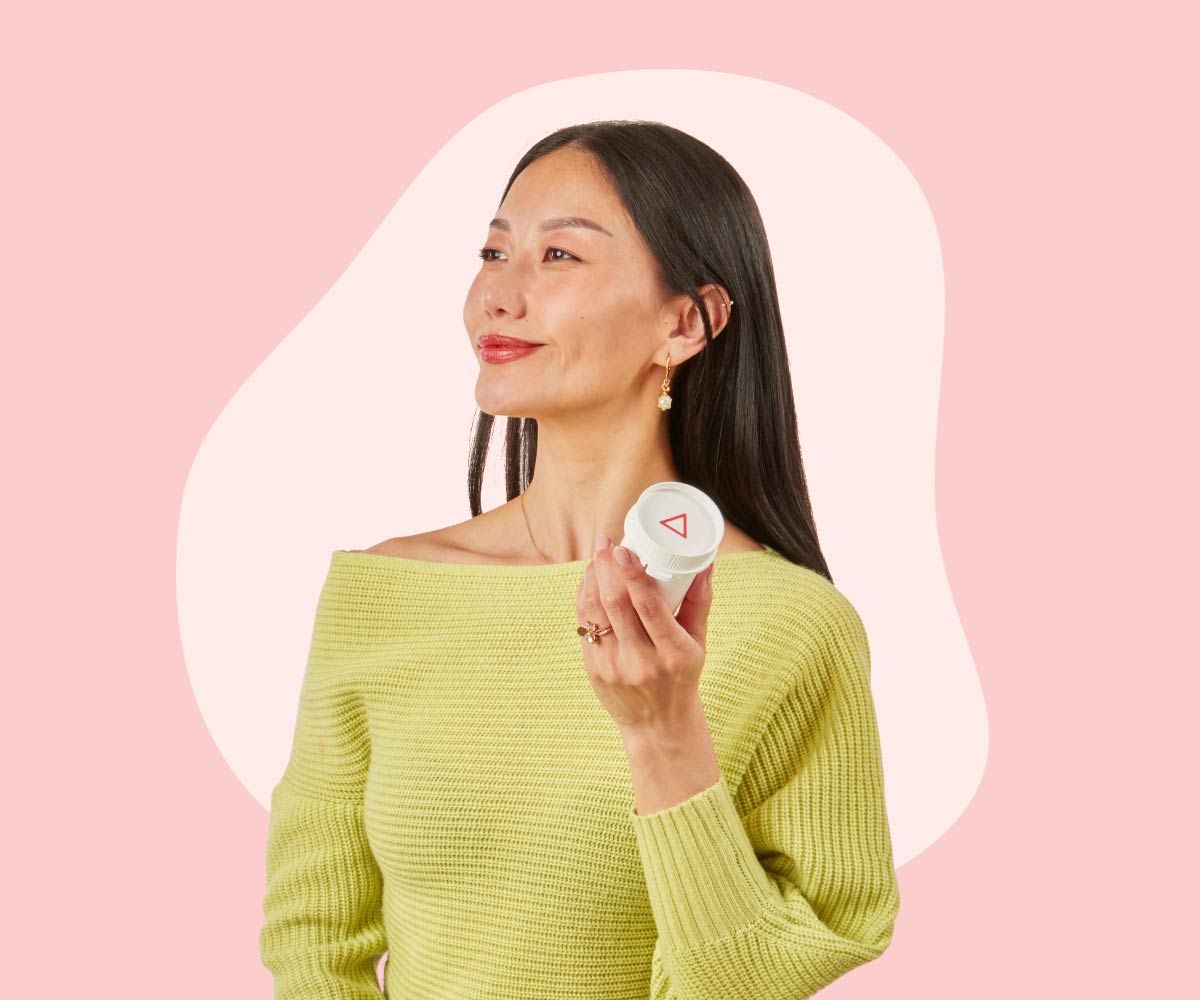 A woman wearing a bright green sweater looks proud while holding a Wisp pill vial in front of a pink background