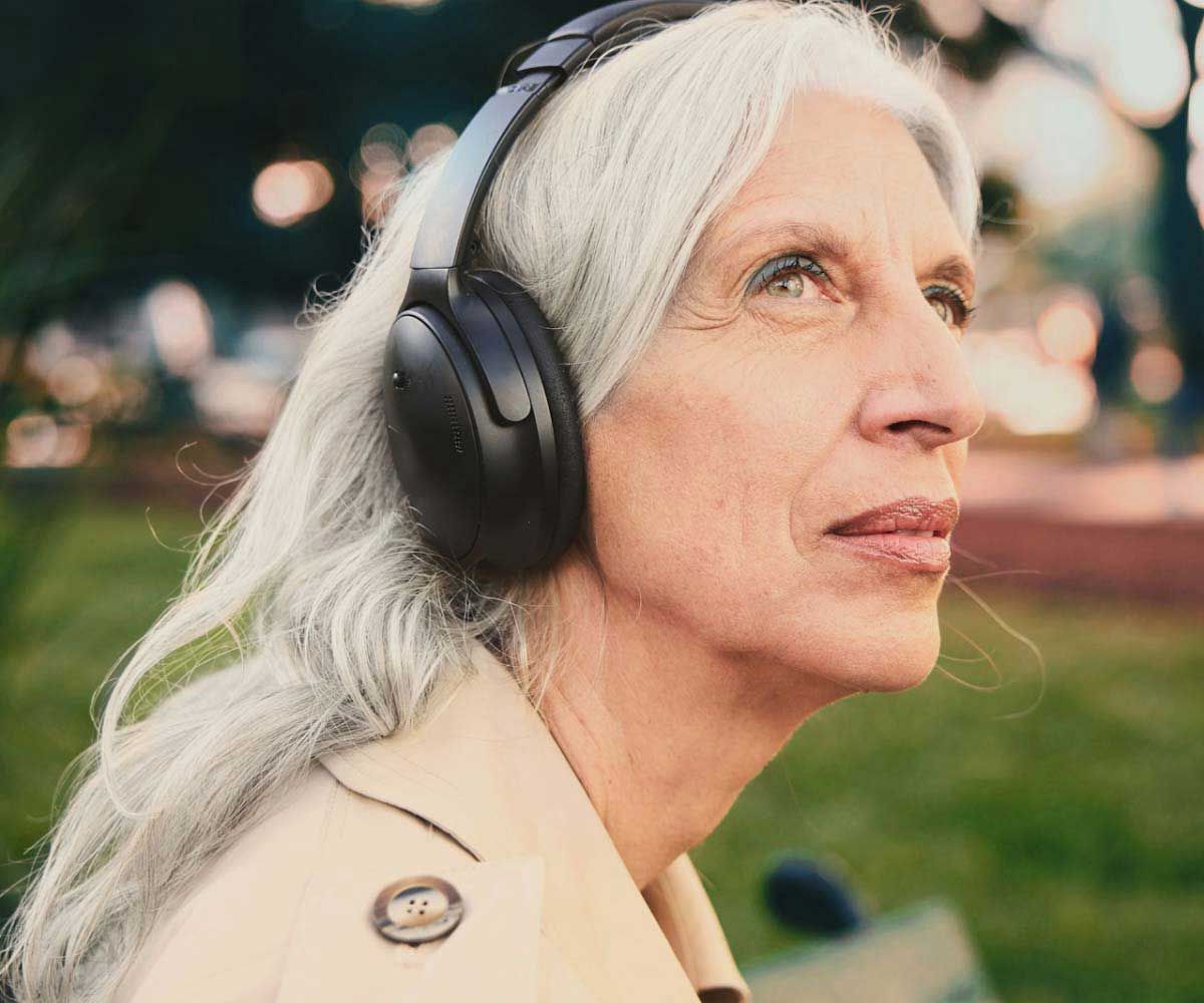 An older woman wearing headphones and a trench coat ouotdoors