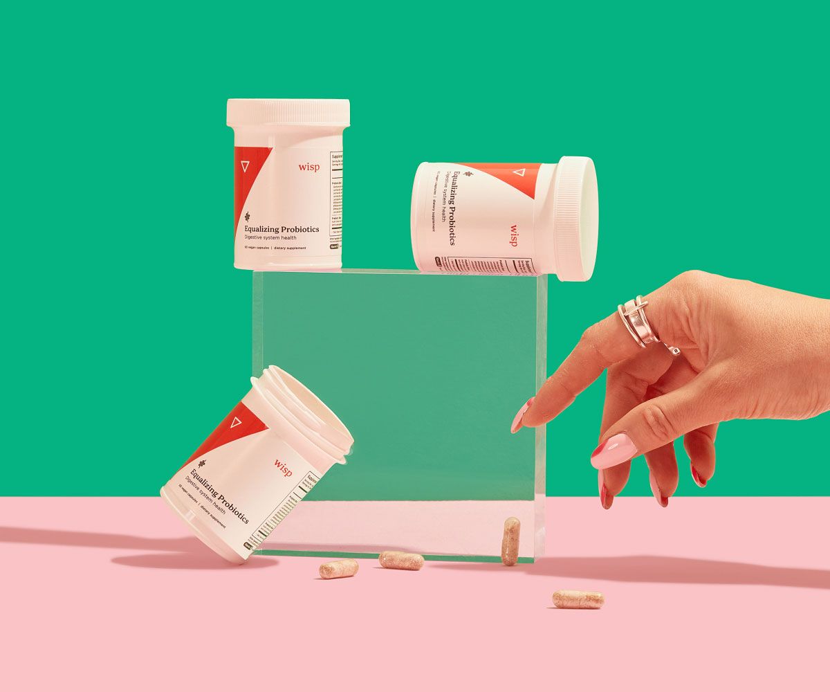 3 bottles of Wisp Equalizing Probiotics balanced on a clear block with pills spilling out and a woman's hand reaching for them on a pink surface with a green background
