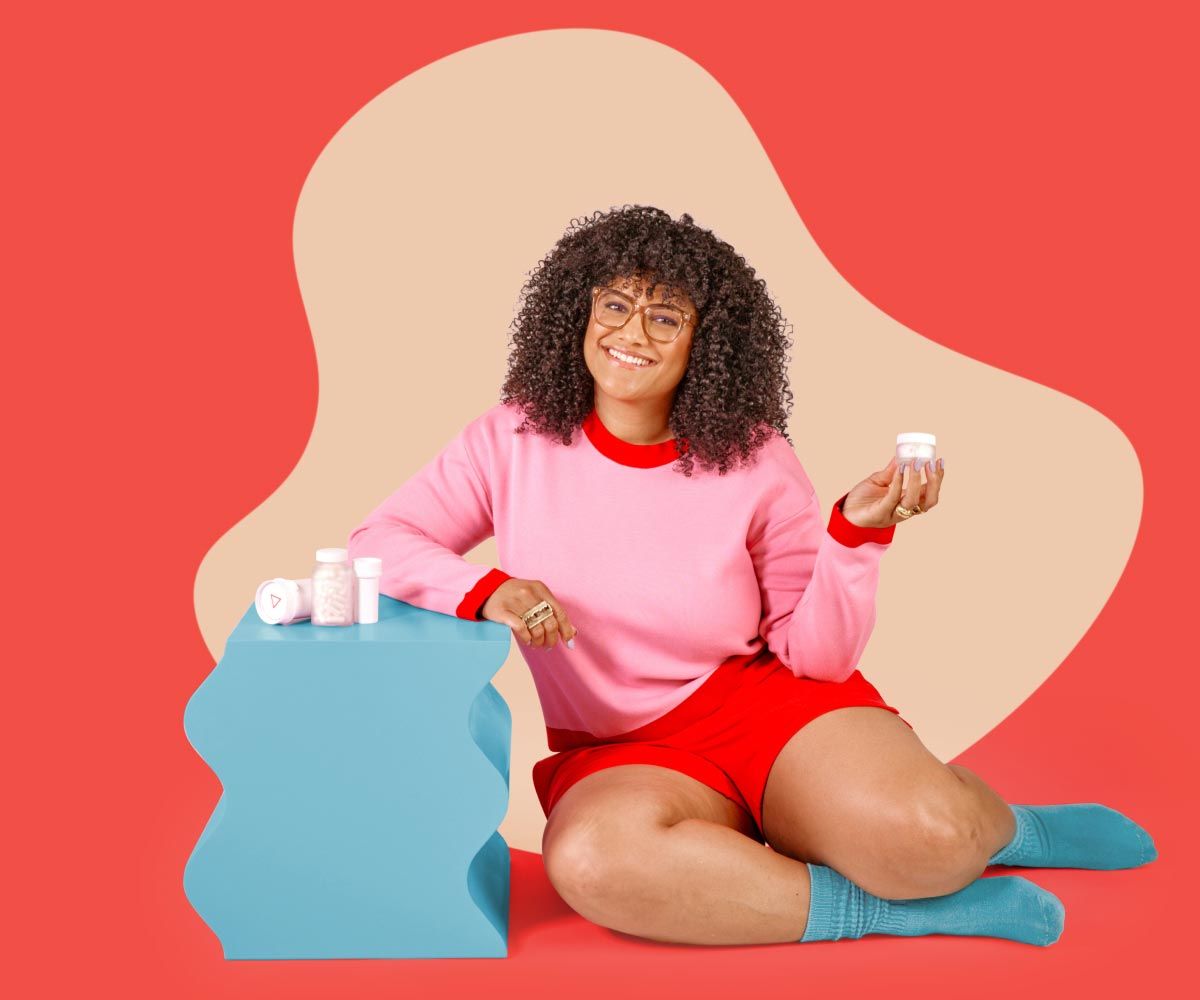 A woman is smiling and wearing a pink and read sweater and shorts with blue socks while sitting on a red surface next to a blue nightstand with several Wisp medications on it