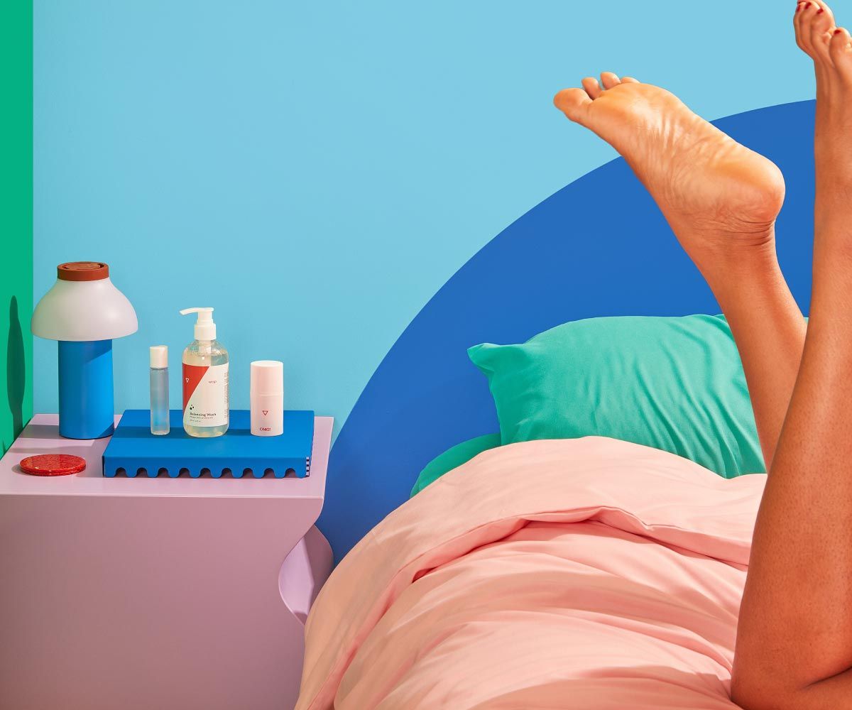 3 Wisp products on a pink nightstand with a blue lamp and next to a bed with colorful bedding and a woman's intertwined legs