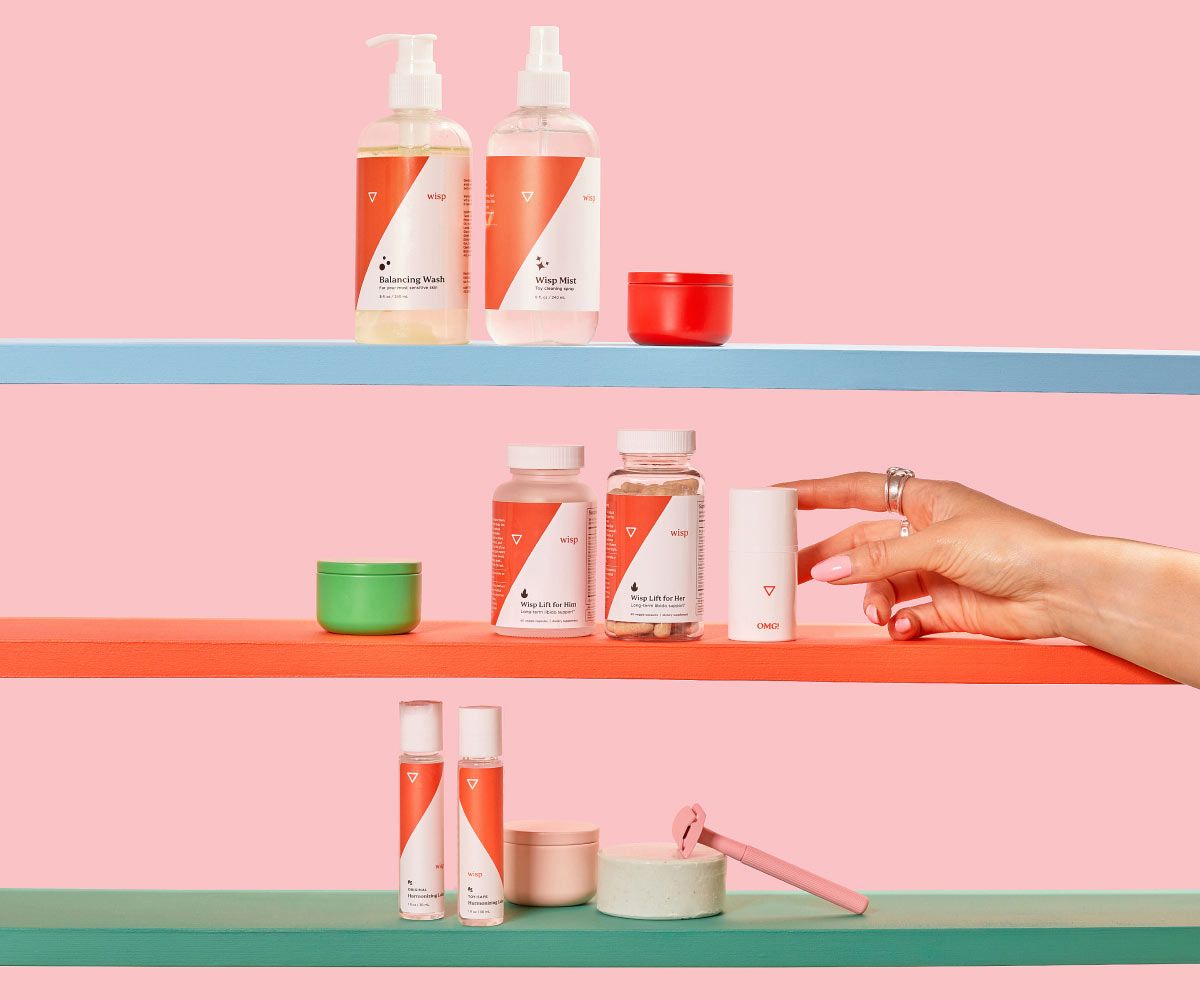 Various Wisp products on colorful shelves with a pink razor and misc containers and a woman's hand reaching for OMG Cream in front of a pink background