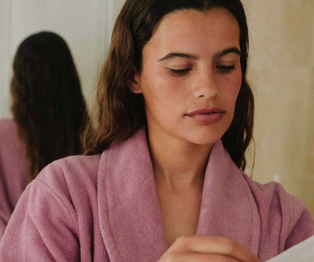 A woman with wavy hair wearing a pink robe is opening a sheet mask