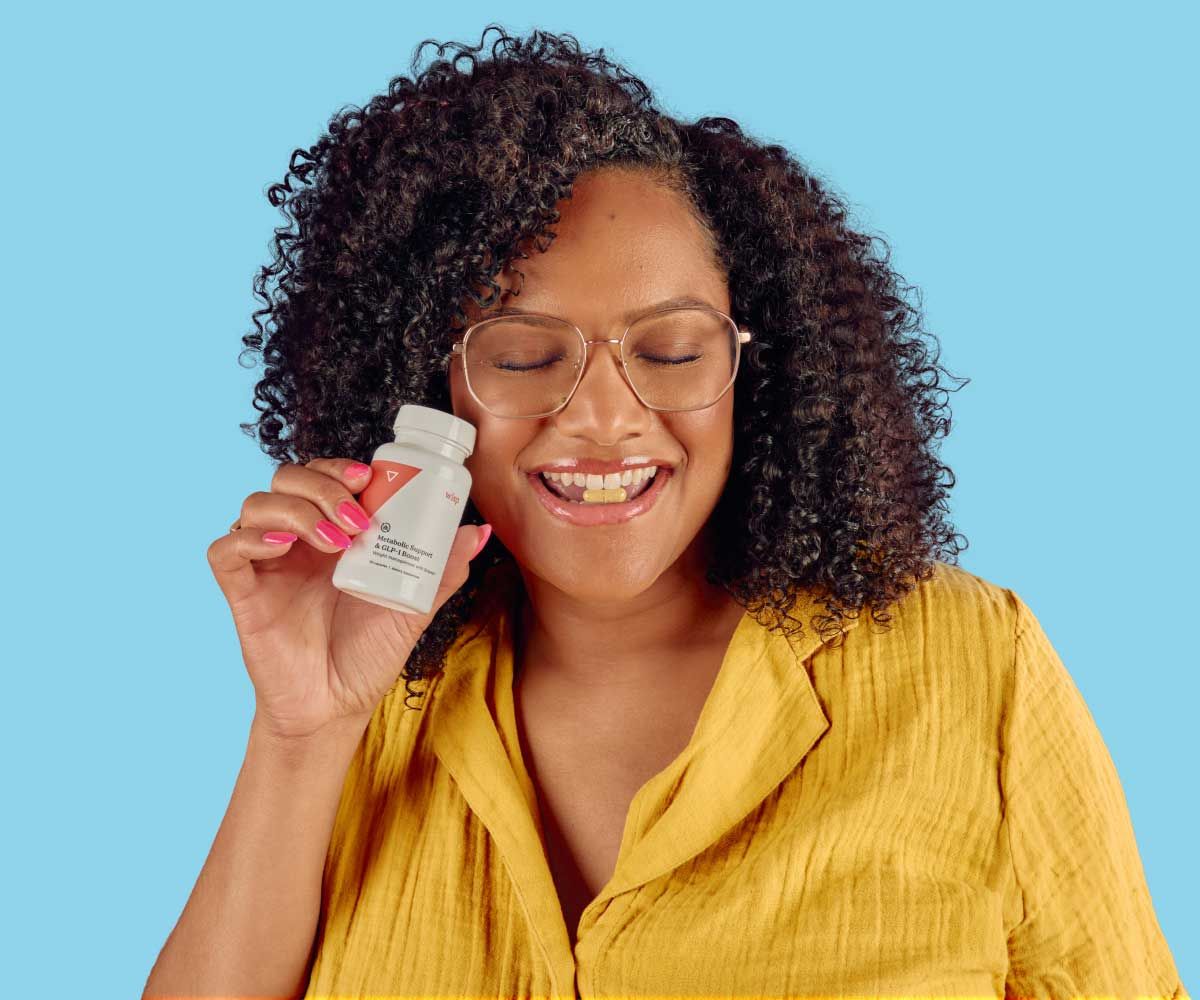 A woman wearing a yellow shirt is holding a bottle of Wisp supplements with a pill in her mouth