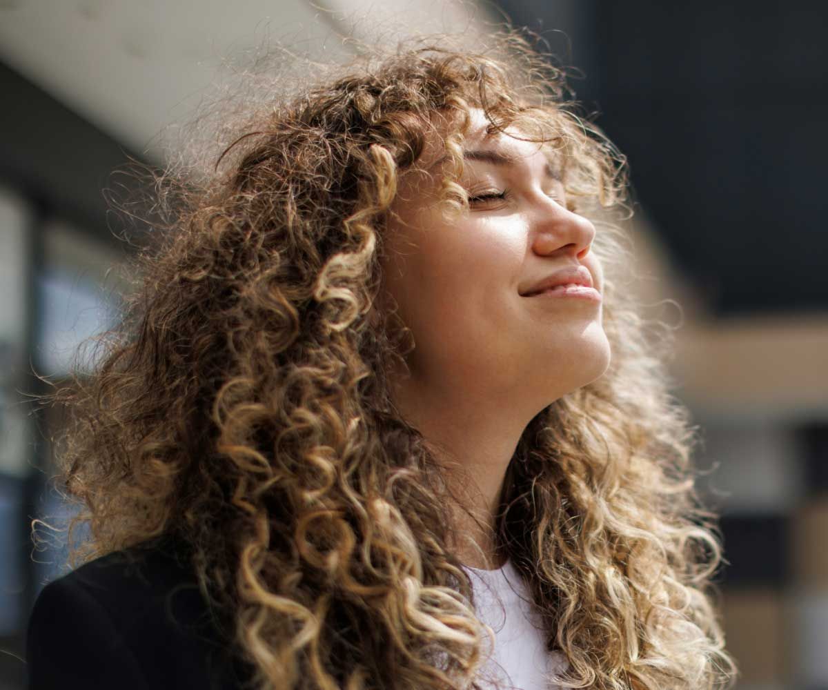 A happy, peaceful woman with curly hair has her head turned up to the sky with sunlight on her face