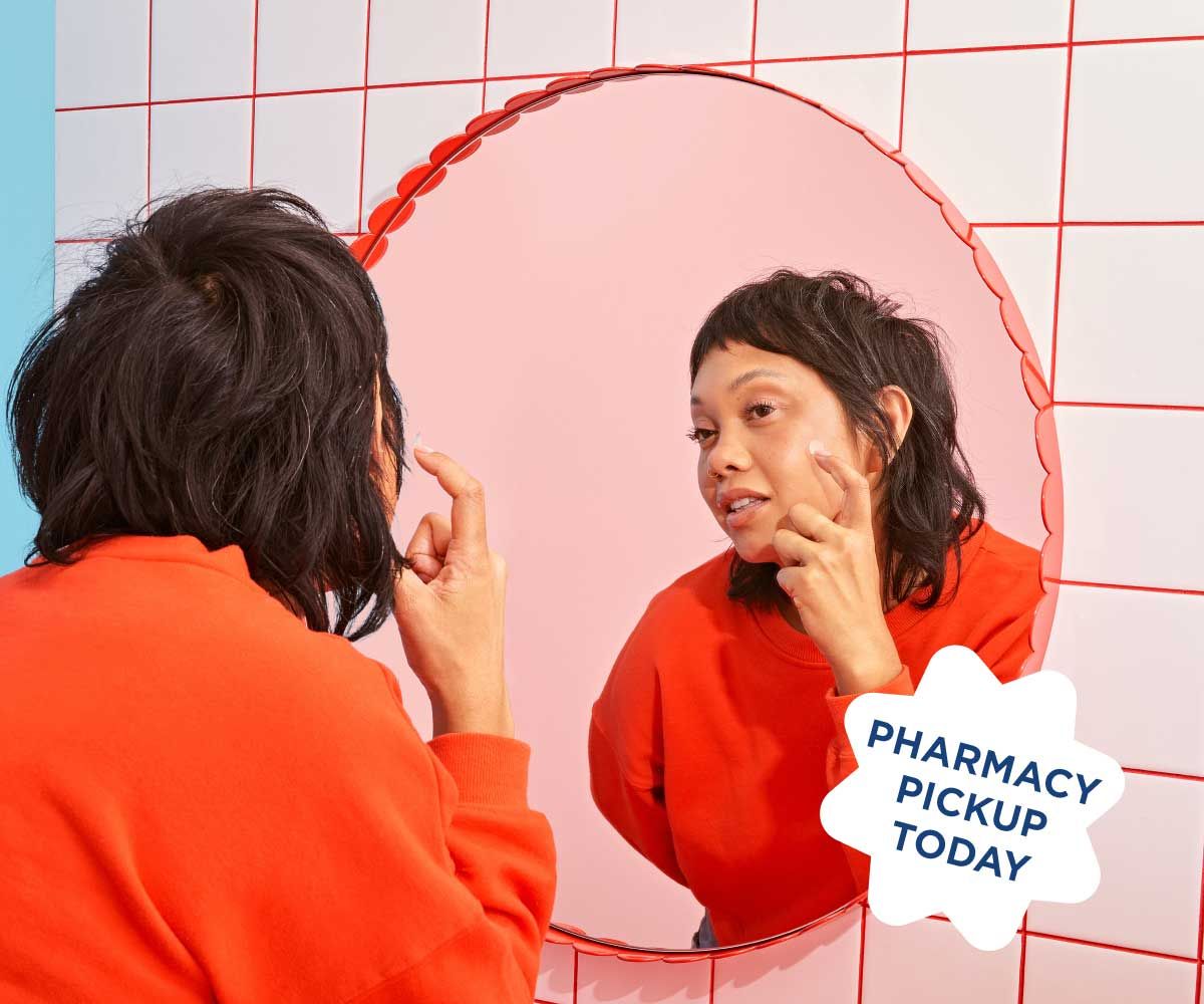 A woman wearing a red sweater is looking into a mirror while putting a hydrocolloid patch on her face