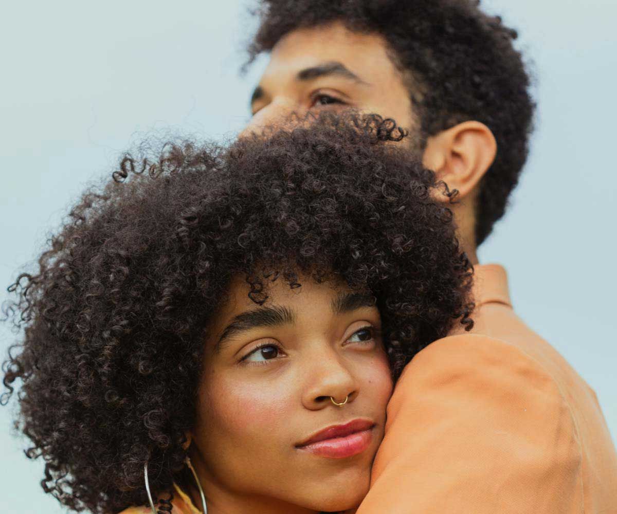 A man and woman with curly hair hugging with a light blue background