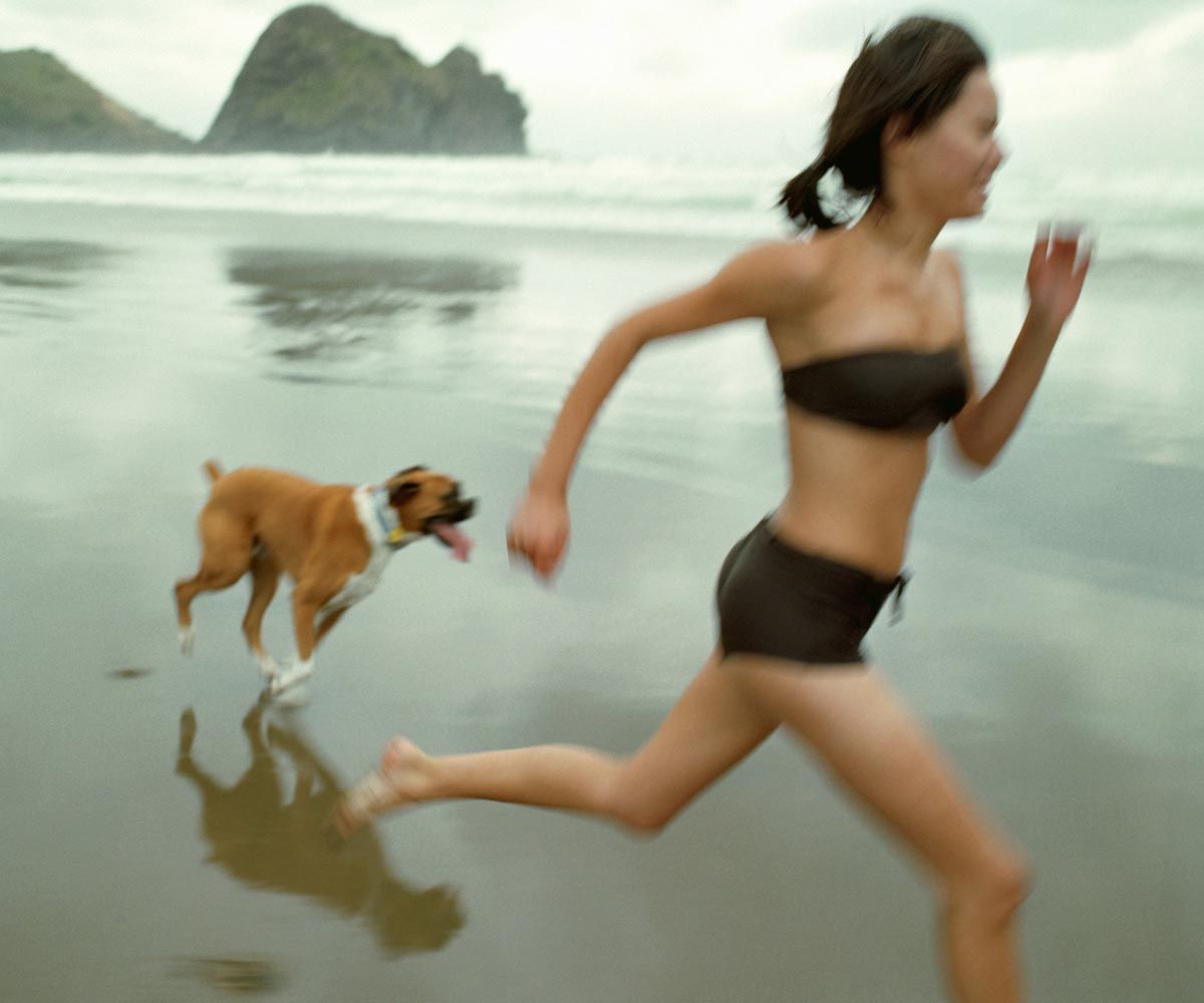 A woman wearing a black swimsuit is running on the beach with her dog