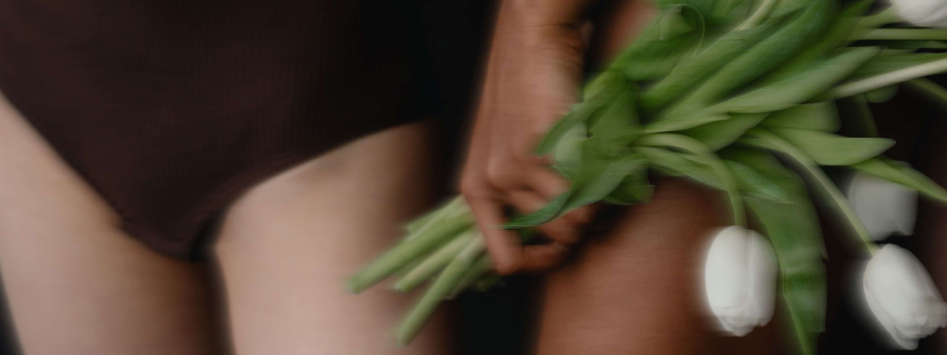 Close-up of a bare leg and hip, with a hand holding white tulips against the thigh; soft, blurred, artistic style.