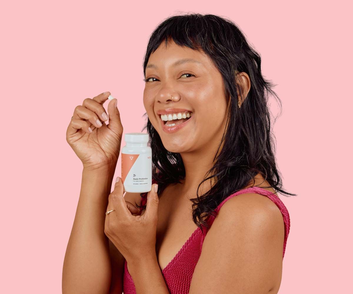 A happy woman wearing a red tank topis holding a pill and a Wisp bottle in front of a pink background