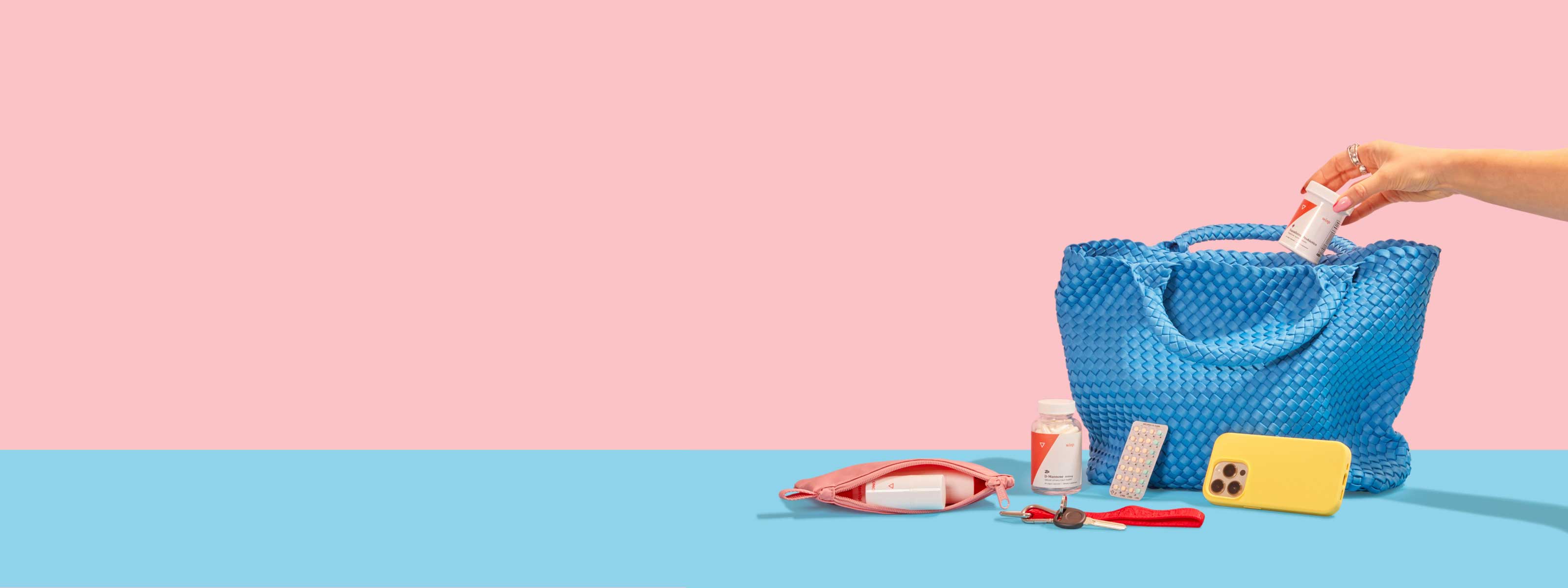 A person's hand is putting Wisp Equalizing Probiotics into a blue handbag with other Wisp Medication, a mobile phone, keys and a travel pouch on a blue surface with a pink background