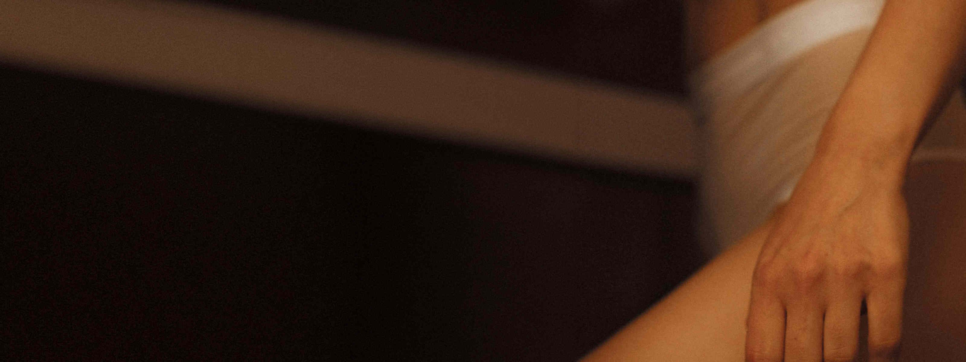 Close-up of a bare arm, softly lit by a diagonal beam of warm light, in low, shadowy lighting.